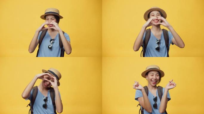 快乐的青春美丽的亚洲女人旅行者戴着帽子和背包，展示迷你心与双手爱情标志微笑看着黄色背景的相机。