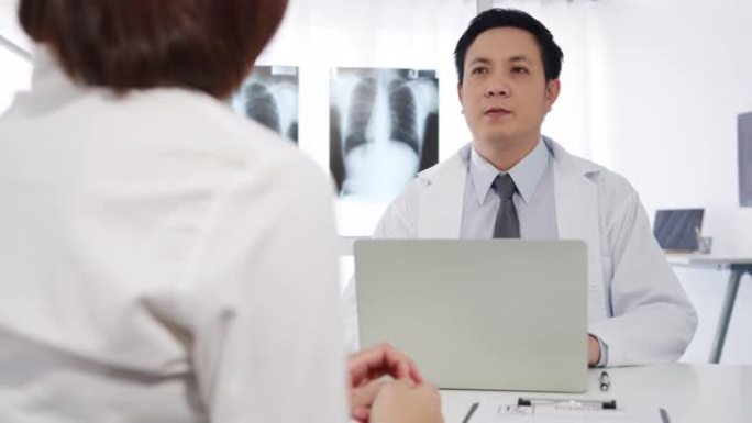 严重的亚洲男性医生使用电脑笔记本电脑正在发表重大新闻谈话与女性患者坐在医院办公室的办公桌前讨论结果或