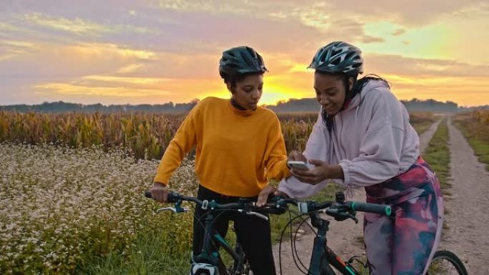 手持慢动作的两名成年女性看着智能手机，在草地土路上骑自行车，背景是日落