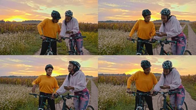 手持慢动作的两名成年女性看着智能手机，在草地土路上骑自行车，背景是日落