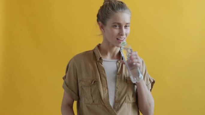 黄色背景上可重复使用的玻璃瓶中的年轻女性饮用水的肖像