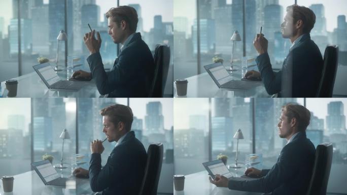 自信的商人穿着西装，坐在现代办公室的办公桌前，使用笔记本电脑，在窗户旁边，可以看到摩天大楼。成功的财