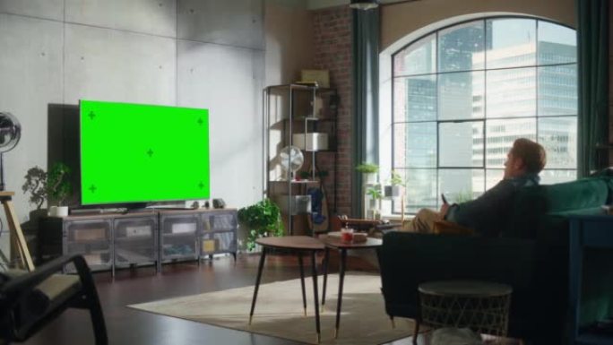 年轻英俊的男人花时间在家里，坐在沙发上，在时尚的阁楼公寓里打开带有绿屏模拟显示的电视。男人在家流电影