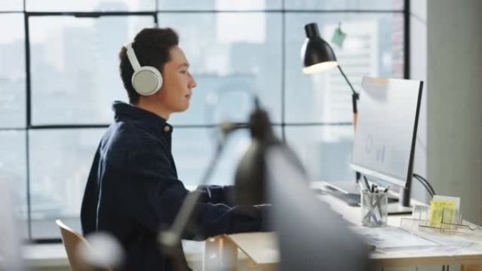 在明亮的现代办公室里，一个亚洲人在使用电脑和耳机时微笑的肖像。传播协调员在收听播客的同时计划一个项目