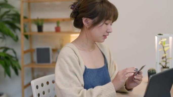 年轻的亚洲女性坐在家里客厅的沙发上，用智能手机在线购物，用信用卡进行移动支付。