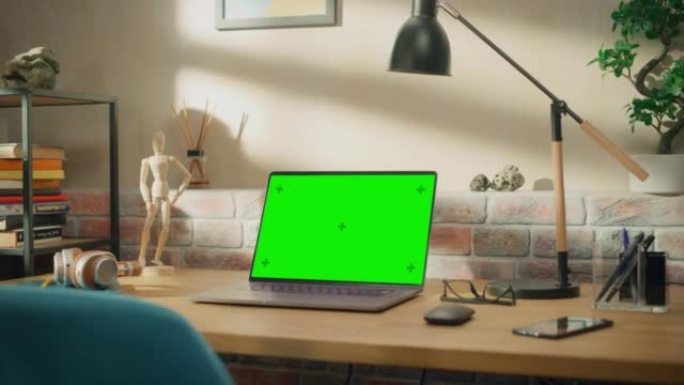 笔记本电脑站在木桌上，绿屏Chromakey模拟显示器。舒适的空阁楼公寓，桌子上有一盏灯、笔记本和智