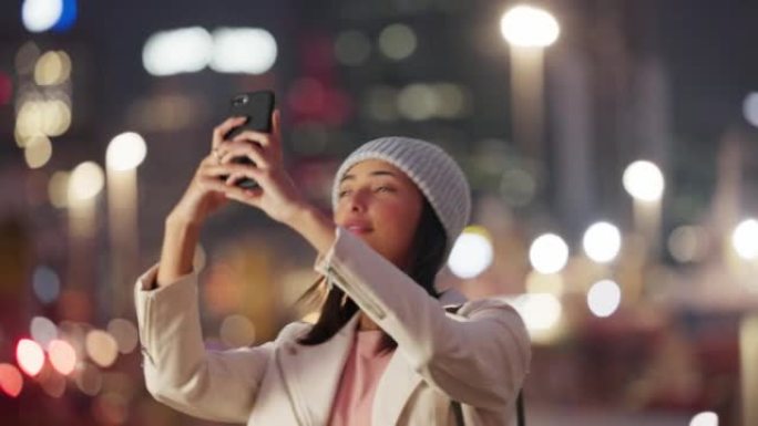 时髦的夜生活视频记录器拿着她的手机，给城市的灯光拍照。年轻女子欣赏美丽的城市小镇，制作了一段充满活力