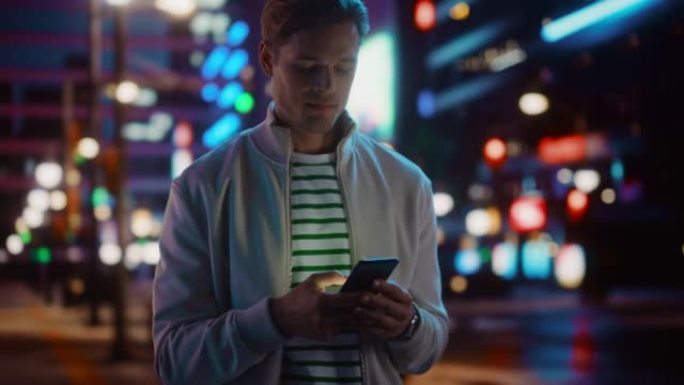 英俊的男人使用智能手机漫步在充满霓虹灯色彩和娱乐的夜城。时尚的年轻人使用手机，在社交媒体上发帖，在线