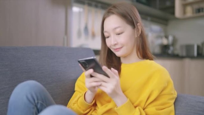 微笑的肖像美丽的亚洲年轻女子年龄36岁，穿着五颜六色的黄色衬衫，用手机看着相机，早上在家坐在沙发上放
