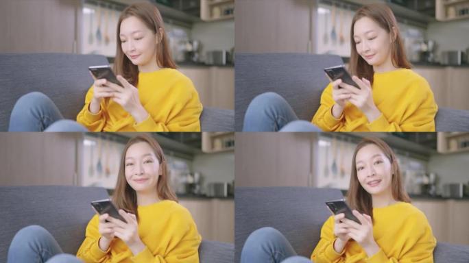 微笑的肖像美丽的亚洲年轻女子年龄36岁，穿着五颜六色的黄色衬衫，用手机看着相机，早上在家坐在沙发上放