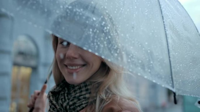 在城市里，白天下雨时，特写肖像女人在伞下，笑容灿烂。时尚女性享受户外城市生活背景下的秋天滴。漂亮的眼