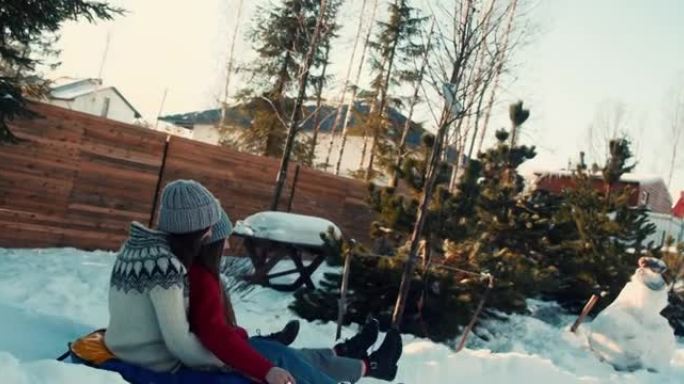 两个迷人的快乐年轻女人在雪坡上向阳光明媚的房子微笑着雪橇。寒假休闲慢动作。