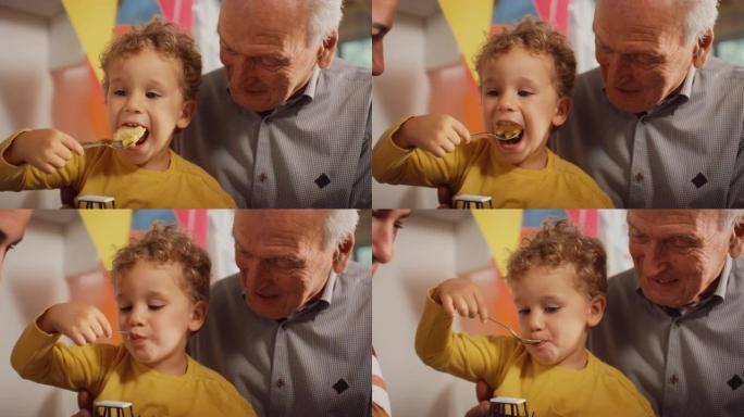 一个可爱的小男孩坐在他爷爷的腿上，吃着美味的生日蛋糕。男孩蹒跚学步的孩子在派对上玩了很长时间后，正在