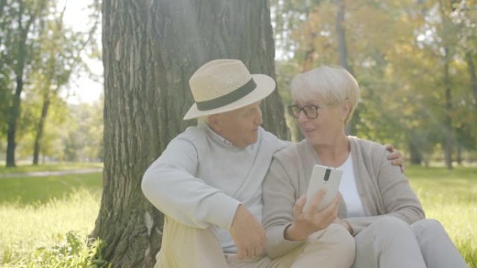 开朗的老年人使用智能手机讨论坐在绿色公园草坪上的媒体内容