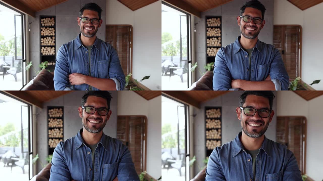 英俊的拉丁美洲男人在家里对着镜头微笑，双臂交叉戴着眼镜