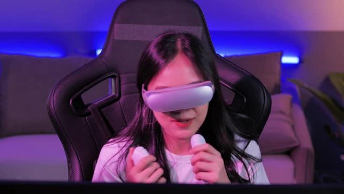 亚洲少女头戴虚拟现实头戴式耳机，一边坐在办公桌前，一边用操纵杆/控制器打手势。夜间在彩色紫外线照明客