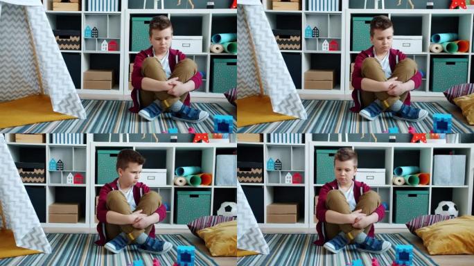 不幸的男孩坐在装满玩具的房间里的地板上，在家中感到无聊和沮丧