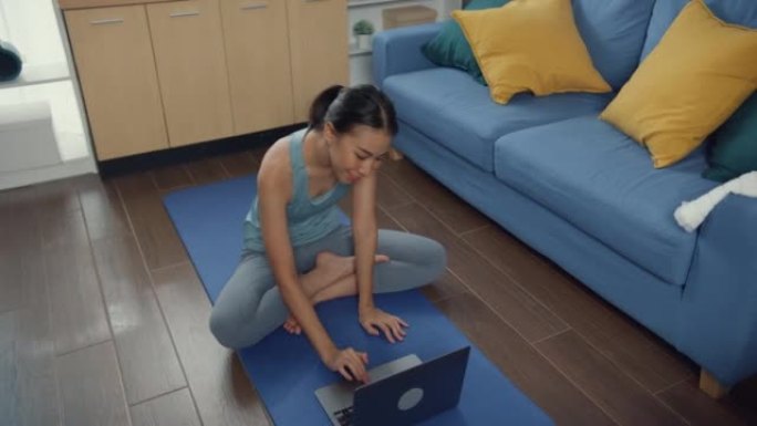 年轻迷人的亚洲女性在运动服网站在地板上的垫子上观看健身在线视频在笔记本电脑上练习瑜伽在家里的客厅初学