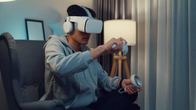 年轻的亚洲男子游戏玩家穿着灰色连帽衫，带虚拟现实控制器耳机保持操纵杆控制器创造3D艺术晚上坐在沙发客