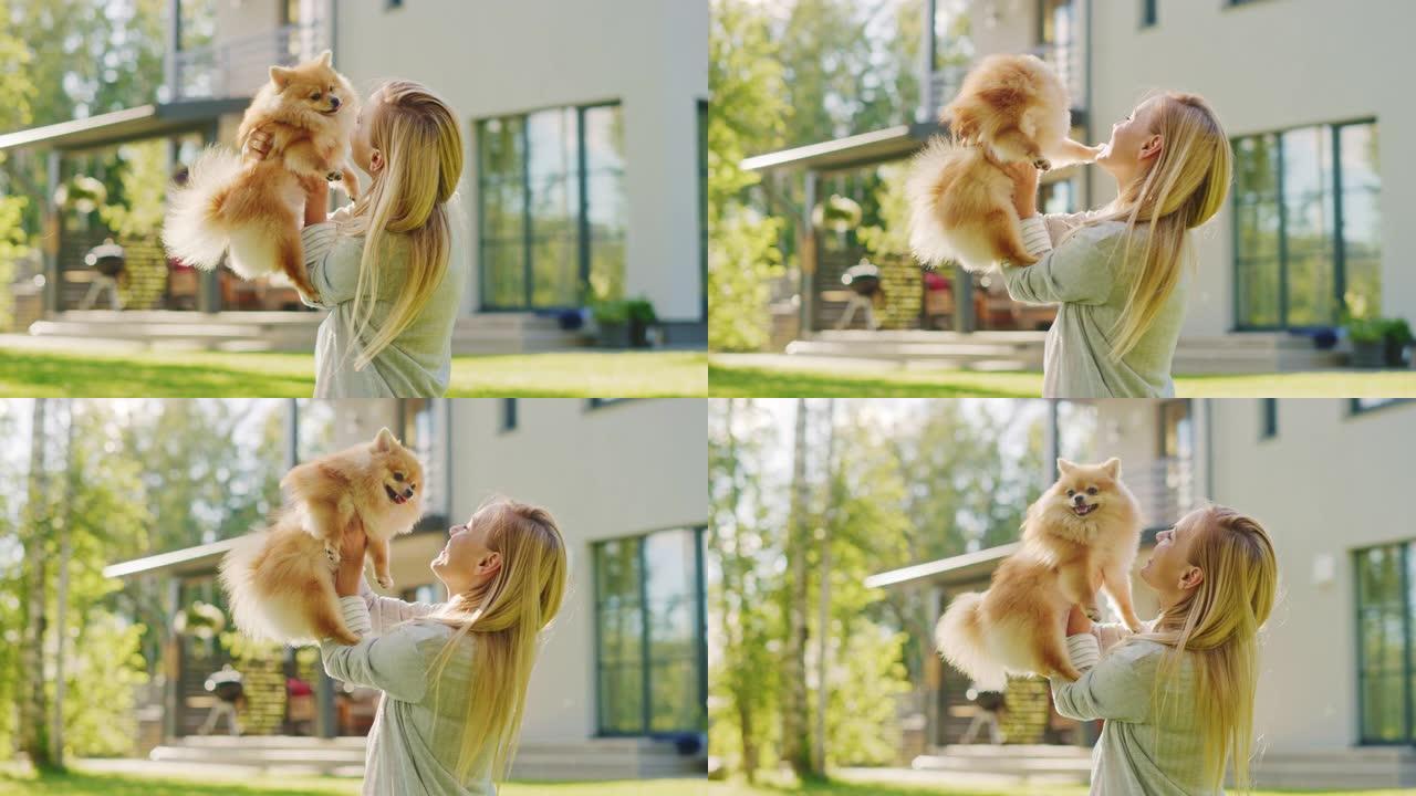 年轻漂亮的女人和她可爱的小博美犬玩耍，抱着她，爱抚，拥抱，几乎亲吻。华丽的晴天，在田园诗般的房子草坪