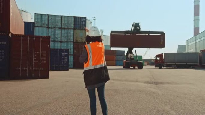女工业工程师，双向收音机，戴着白色安全帽，橙色高能见度背心，为物流作业码头的集装箱搬运工指明卸货位置