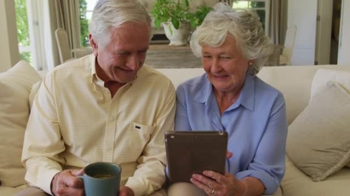 白人高级夫妇一起坐在家里的沙发上，一边微笑着使用数字平板电脑