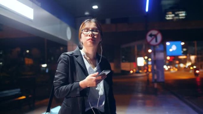 年轻女子带着智能手机在晚上的公共汽车站等候