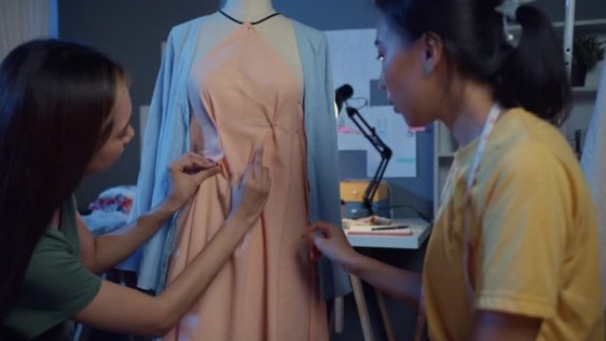 两位专业的亚洲开朗女士时装设计师团队合作，休闲加班，分享想法，晚上在商店假人身上创造漂亮的衣服。女裁