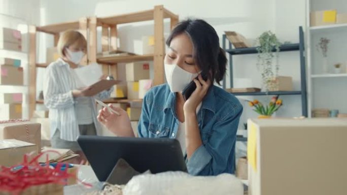 年轻的亚洲女商人戴着口罩，使用手机接听订单，并在家庭办公室检查库存工作中的产品。