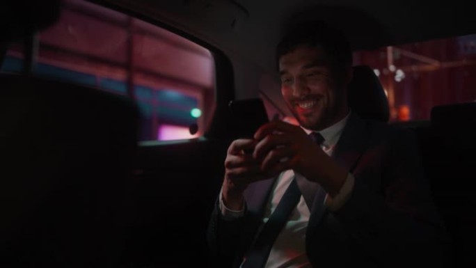 穿着西装的快乐英俊商人晚上坐在汽车后座从办公室通勤。企业家在城市街道上使用智能手机，并在霓虹灯标志下
