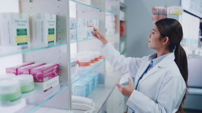 药房药店: 美丽勤奋的亚洲药剂师穿着白大褂的肖像在柜台后面的架子上布置药包，药盒，维生素和补充药丸