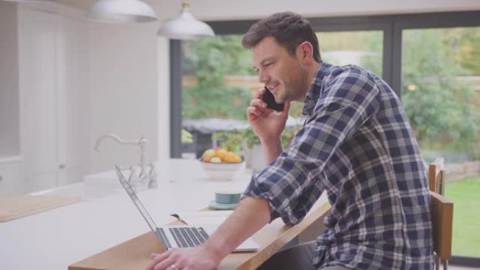 男子在家工作时在厨房柜台上使用笔记本电脑，同时在手机上聊天