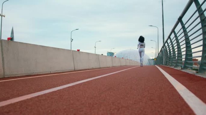 一个穿着白色运动服的健美卷发女跑步者正在桥上慢跑，我们从后面看到她的慢动作
