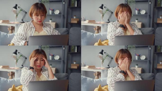 自由亚洲女士坐在沙发上，带着笔记本电脑在线学习，在家里的客厅感到头痛。