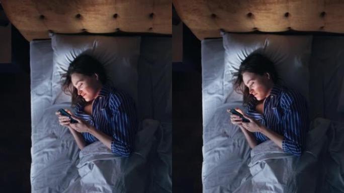 顶视图垂直屏幕卧室公寓: 美丽的年轻女子晚上在床上使用智能手机。微笑的女孩浏览社交媒体，在手机上进行