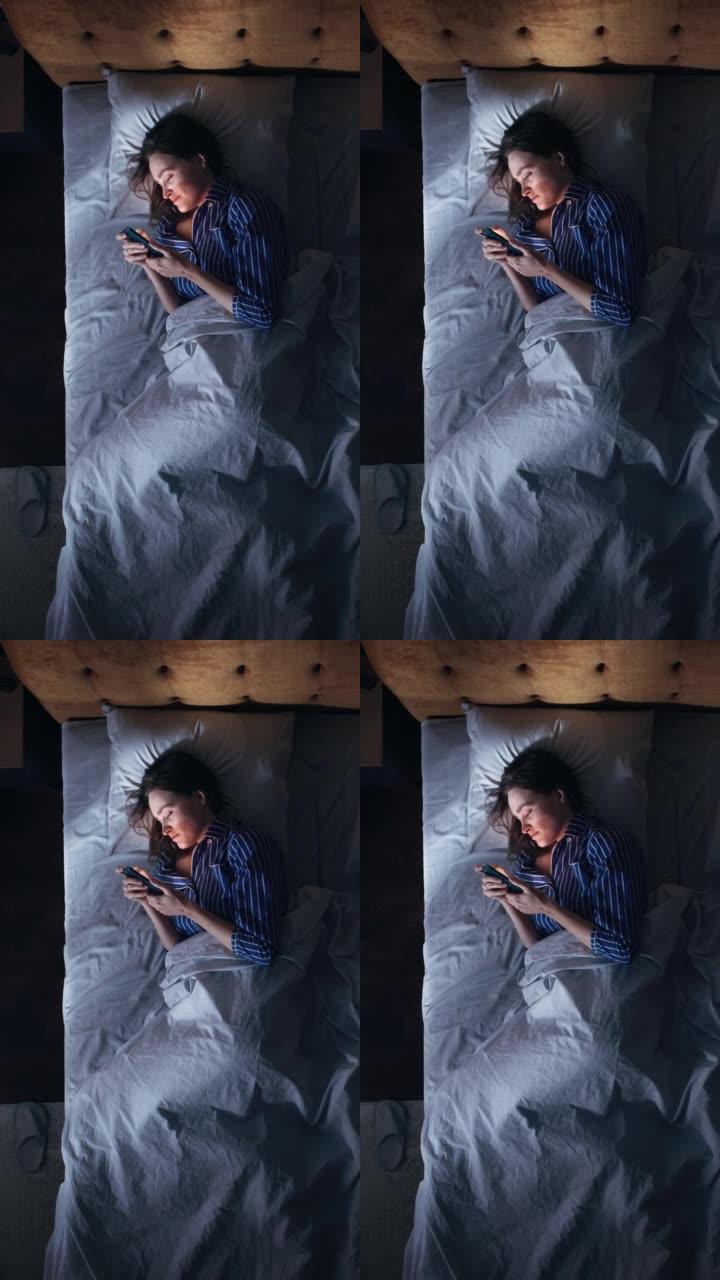 顶视图垂直屏幕卧室公寓: 美丽的年轻女子晚上在床上使用智能手机。微笑的女孩浏览社交媒体，在手机上进行