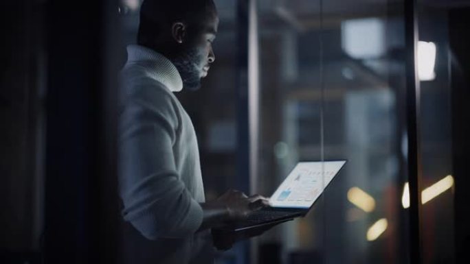 英俊的黑人非洲裔美国男性站在一家创意机构的玻璃墙后面的会议室里，拿着笔记本电脑。项目经理穿着白色套头