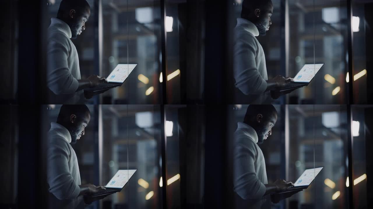 英俊的黑人非洲裔美国男性站在一家创意机构的玻璃墙后面的会议室里，拿着笔记本电脑。项目经理穿着白色套头