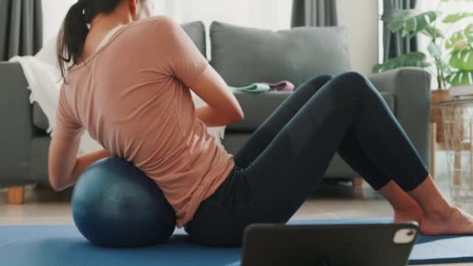 穿着运动服的年轻迷人亚洲女性的特写镜头，在家里的客厅用健身球在平板电脑上观看健身在线视频。