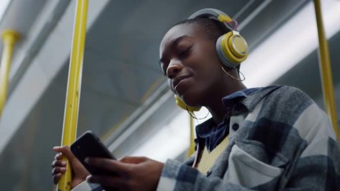 时尚的年轻黑人女性晚上在公共汽车上使用智能手机和耳机。微笑和快乐的非洲裔美国人聊天，发短信，在互联网