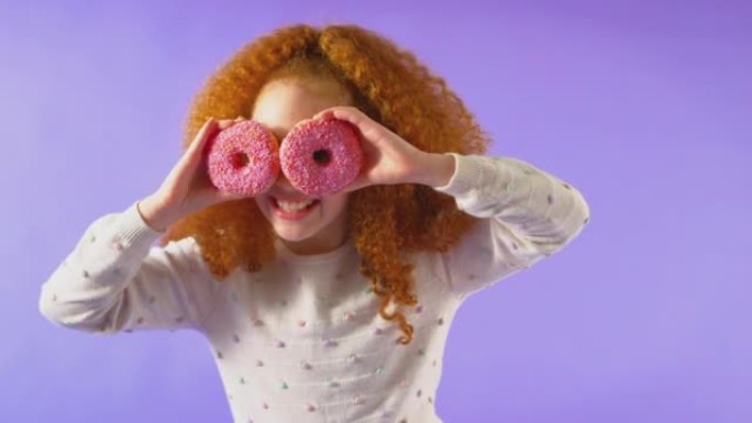 在紫色工作室背景下，红发年轻女孩像眼镜一样在脸前拿着两个甜甜圈的工作室镜头-慢动作拍摄