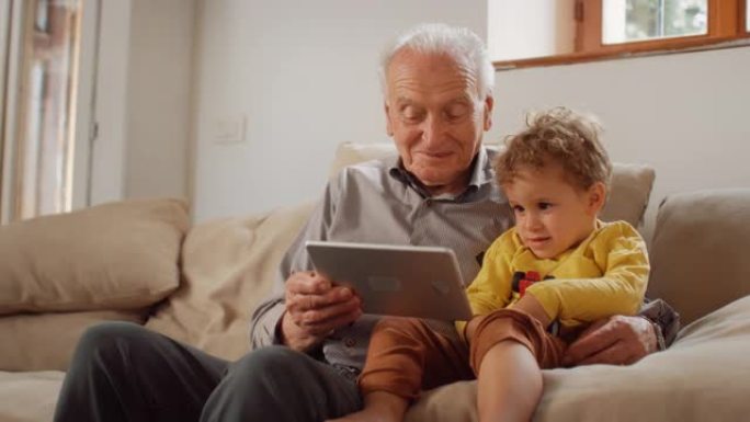 一个老人坐在沙发上与他的小孙子一起使用平板电脑的慢动作肖像。可爱的孩子正在听他的祖父正在使用技术阅读