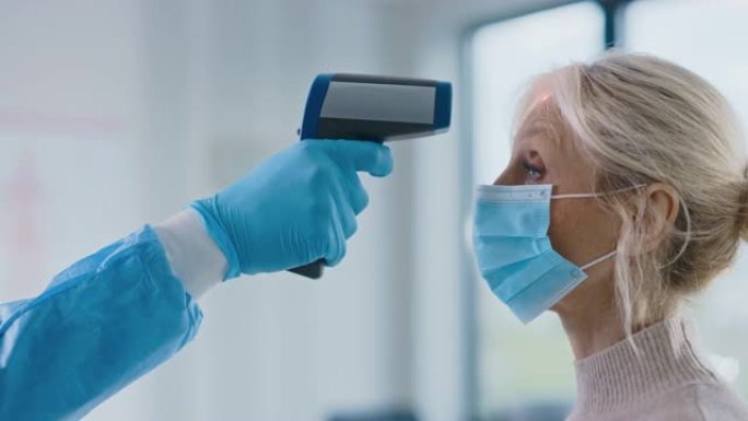 在健康诊所中，一名戴着安全手套的医护人员用红外温度计测量高级女性患者的体温。医生使用免触摸技术来诊断