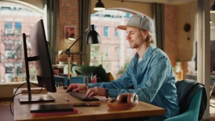年轻英俊的男人在阁楼公寓用台式电脑工作。创意男性查看社交媒体，在家浏览互联网。从大窗户可以看到城市的