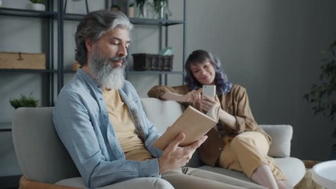 丈夫看书，微笑，而妻子在背景中使用智能手机
