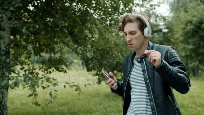 快乐的男人戴着耳机跳舞的慢动作使用智能手机在城市公园散步
