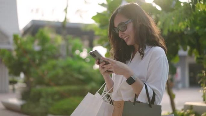 亚洲女子手提购物袋购物后在城市街道用手机上网