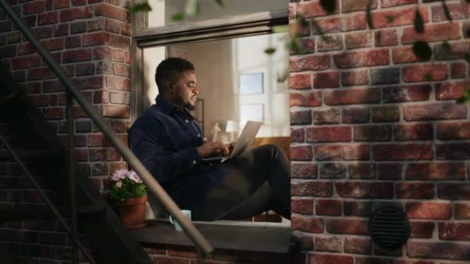 黑人年轻男性坐在窗台上微笑着使用笔记本电脑。非裔美国商人在家工作时享受天气。他正在思考和回复他的商业