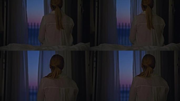 晚上，金发女人坐在酒店房间的床上，看着窗外，背景是大海的阳台