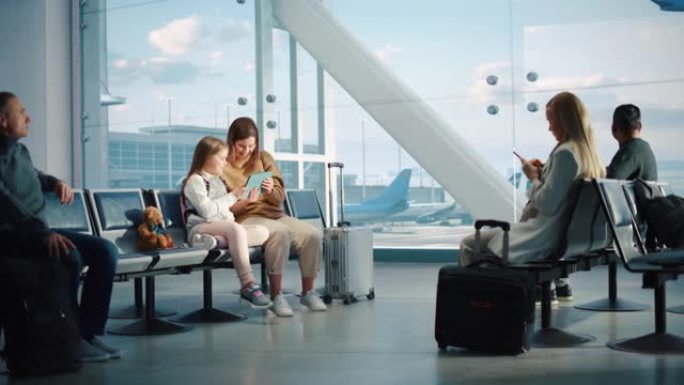机场航站楼: 可爱的母亲和小女儿等待他们的家人度假航班，在数字平板电脑上玩教育游戏。坐在飞机枢纽登机